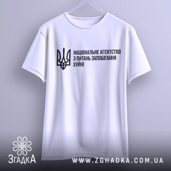 Біла футболка Агентство Запобігання Подарунок, що висить на вішаку, з виразним написом та гербом.