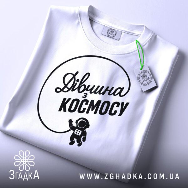 Складена біла футболка з принтом "дівчина з космосу" та етикеткою "Згадка".