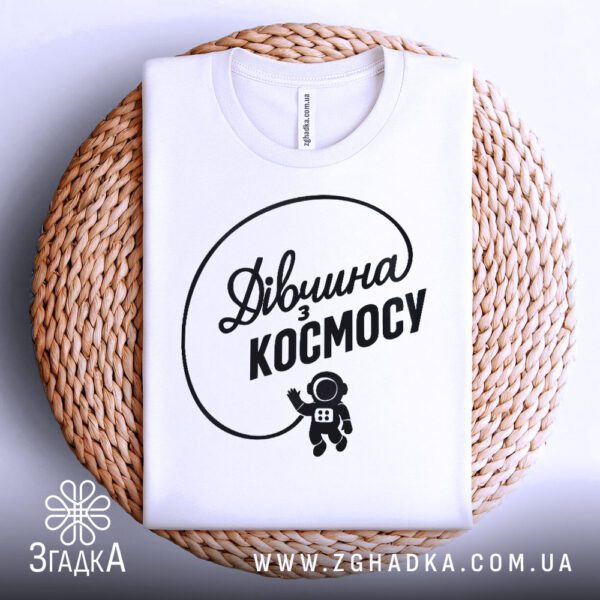 Біла футболка з принтом "дівчина з космосу", акуратно складена на круглій дерев'яній поверхні.