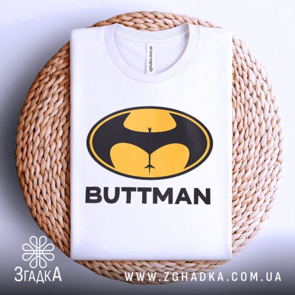 Чорна футболка з принтом "BUTTMAN", акуратно складена на круглій дерев'яній поверхні.