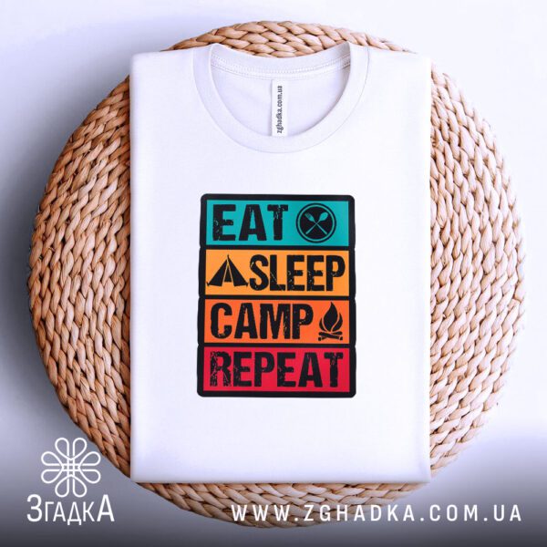Біла футболка з принтом "Eat Sleep Camp Repeat", акуратно складена на круглій дерев'яній поверхні.