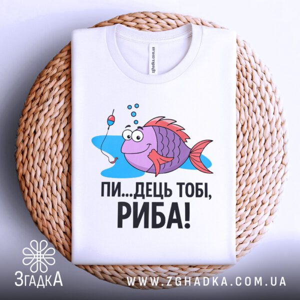 Фронтальний вигляд білої футболки з кольоровим принтом веселої риби-рибалки та написом "ПИ...ДЕЦЬ ТОБІ, РИБА!", розташованої на плетеному круглому підносі.