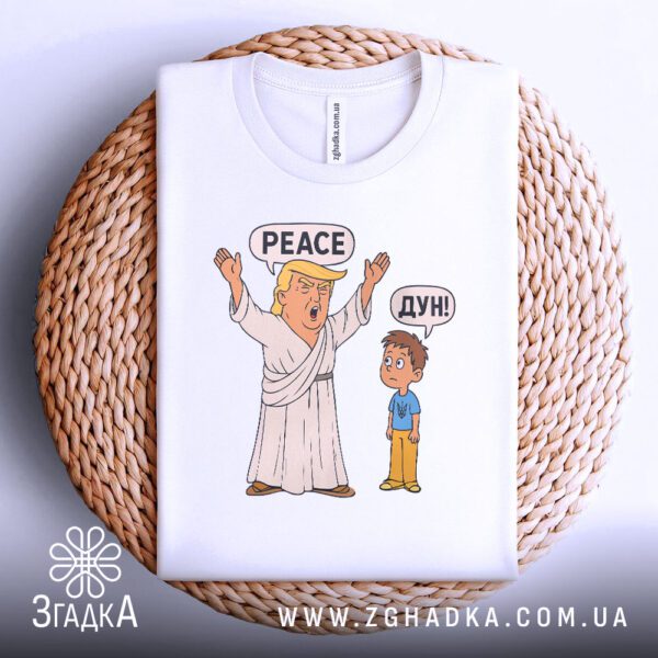 Біла футболка з принтом "Дональд Трамп пародія", акуратно складена на круглій дерев'яній поверхні.