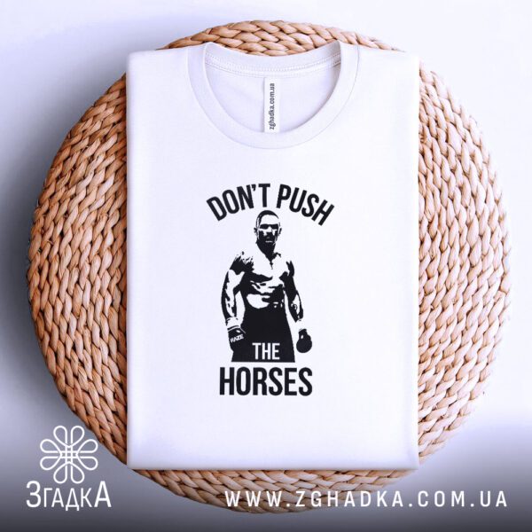 Біла футболка з принтом "don't push horses", акуратно складена на круглій дерев'яній поверхні.