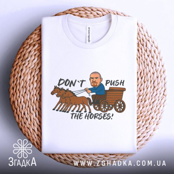 Біла футболка з принтом "don't push карикатура", акуратно складена на круглій дерев'яній поверхні.