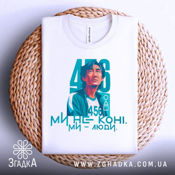 Фронтальний вигляд білої футболки з яскравим принтом головного героя серіалу "Гра в кальмара" під номером 456 та написом "МИ НЕ КОНІ. МИ - ЛЮДИ.", розташованої на плетеному круглому підносі.