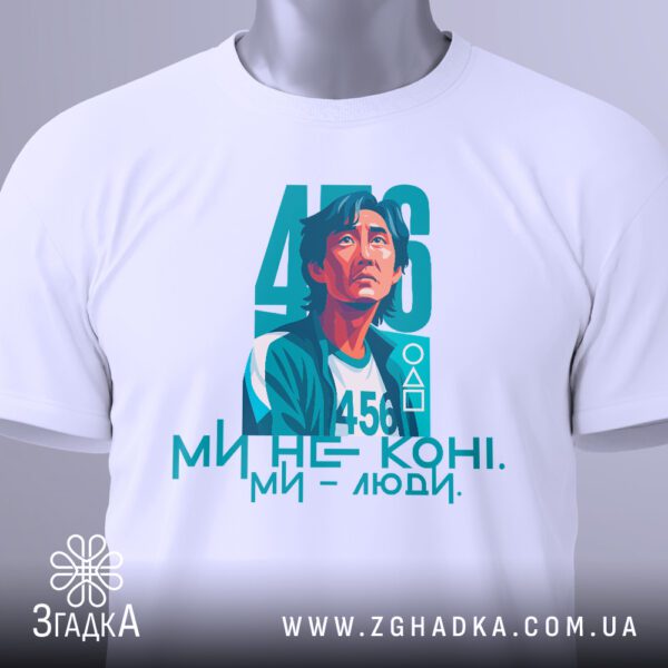 Крупний план яскравого принту головного героя серіалу "Гра в кальмара" під номером 456 та написом "МИ НЕ КОНІ. МИ - ЛЮДИ." на білій футболці.