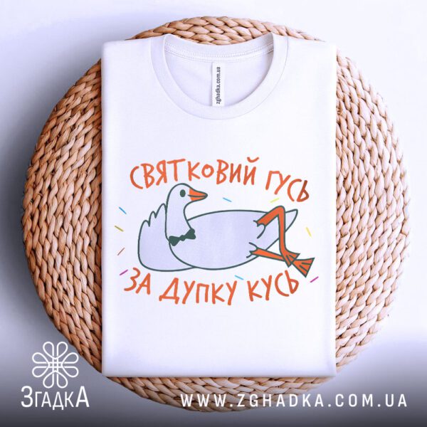 Біла футболка з принтом "гусь дупку кусь", акуратно складена на круглій дерев'яній поверхні.