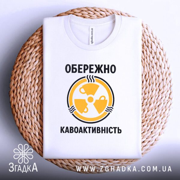 Біла футболка з принтом "кавоактивність з іронією", акуратно складена на круглій дерев'яній поверхні.