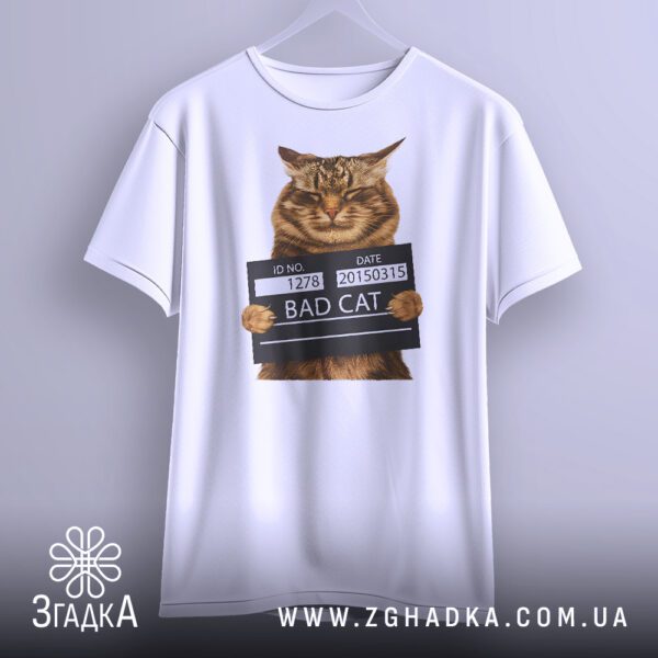 Біла футболка з принтом "кіт bad cat", що висить на білих плічках, демонструючи дизайн.