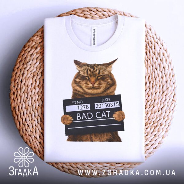 Біла футболка з принтом "кіт bad cat", акуратно складена на круглій дерев'яній поверхні.