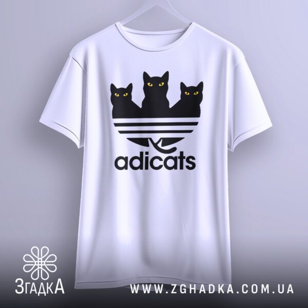 Біла футболка з принтом "кіт пародія Adidas", що висить на білих плічках, демонструючи дизайн.