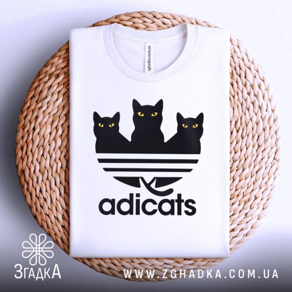 Біла футболка з принтом "кіт пародія Adidas", акуратно складена на круглій дерев'яній поверхні.