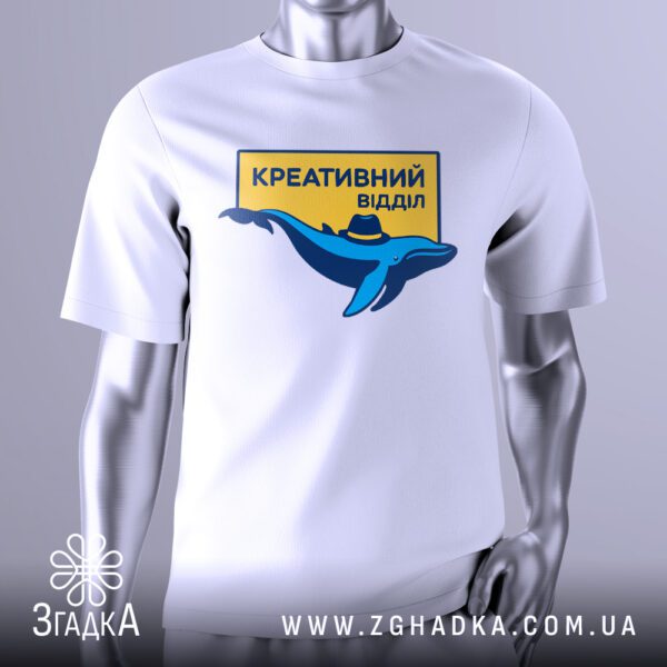 Біла футболка з графічним принтом у центрі. На принті зображено стилізованого синього кита в циліндрі або капелюсі, на жовтому фоні. Над китом і під ним розташований текст українською мовою: "Креативний відділ". Футболка на сірому манекені.