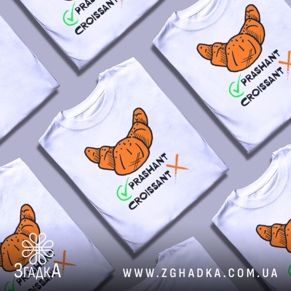 Кілька білих футболок з принтом "круасан став прашант", складені стопкою або розкладені поруч на світлому фоні.