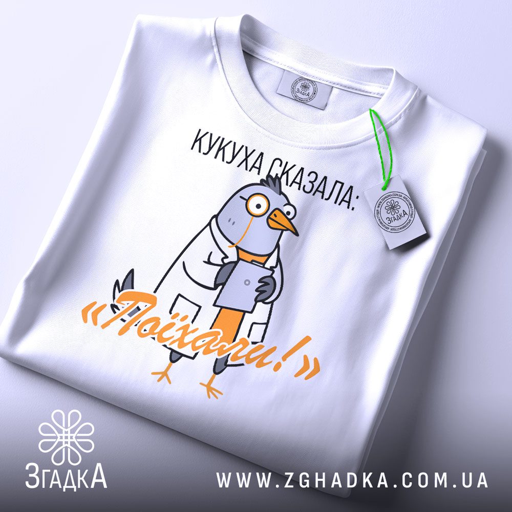 Складена біла футболка з чорним текстовим принтом "Кукуха сказала поїхали" та етикеткою "Згадка".