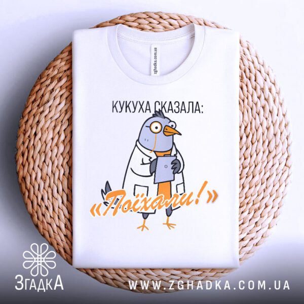 Біла футболка з принтом "Кукуха сказала поїхали", акуратно складена на круглому плетеному підносі.