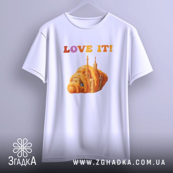 Біла футболка з принтом "love it круасан", що висить на білих плічках, демонструючи дизайн.