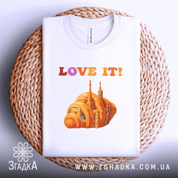 Біла футболка з принтом "love it круасан", акуратно складена на круглій дерев'яній поверхні.