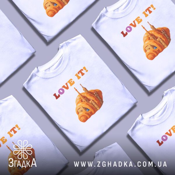 Кілька білих футболок з принтом "love it круасан", складені стопкою або розкладені поруч на світлому фоні.