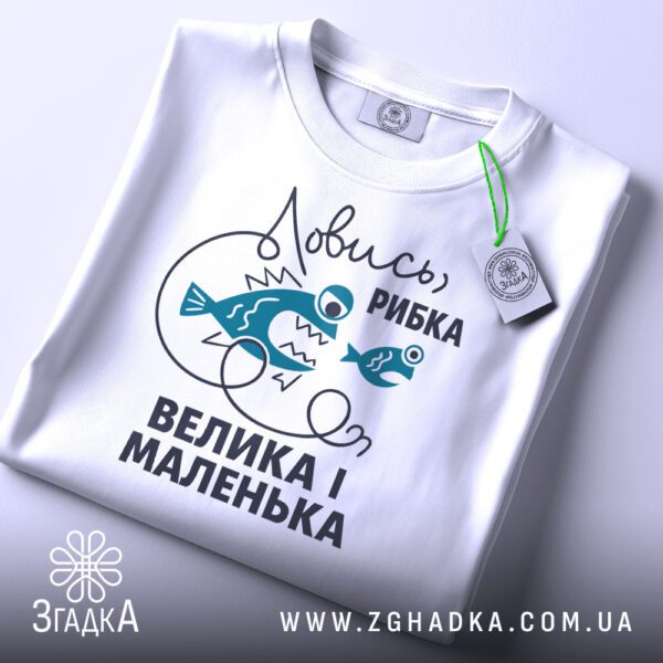 Складена біла футболка з принтом "Ловись рибка велика-велика" та етикеткою "Згадка".