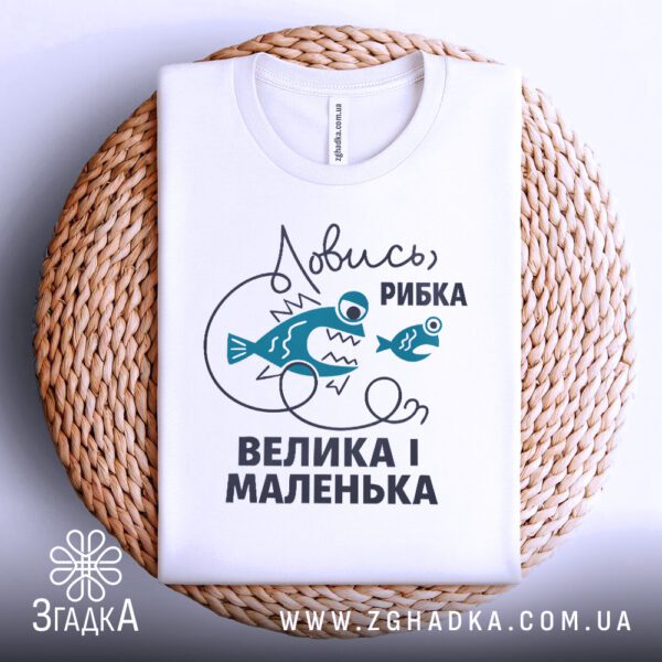 Фронтальний вигляд білої футболки з принтом "Ловись рибка велика-велика", розташованої на плетеному круглому підносі.