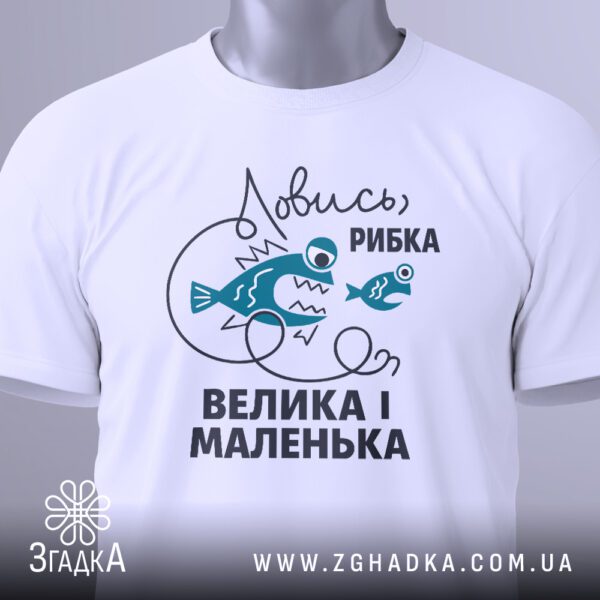 Крупний план кольорового принту чоловічка, що ловить велику зелену рибу, та напису "Ловись рибка велика-велика" на білій футболці.