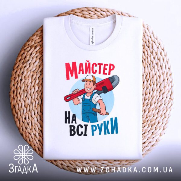 Біла футболка з принтом "Майстер на всі руки", акуратно складена на круглому плетеному підносі.