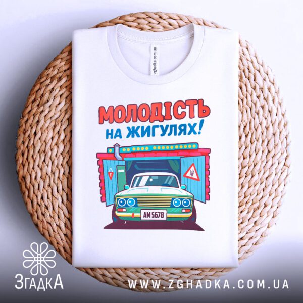 Біла футболка з принтом "молодість на жигулях", акуратно складена на круглій дерев'яній поверхні.