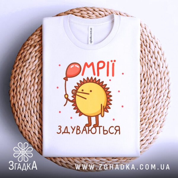 Біла футболка з принтом "мрії здуваються їжак", акуратно складена на круглій дерев'яній поверхні.