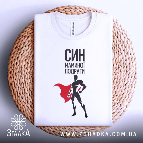 Біла футболка з принтом "СИН МАМИНОЇ ПОДРУГИ", акуратно складена на круглій дерев'яній поверхні.