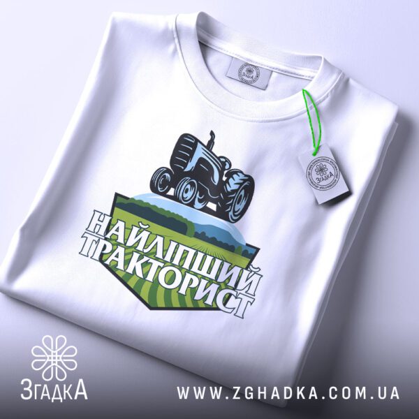 Складена біла футболка Найліпший Тракторист з яскравим принтом та етикеткою згадка.