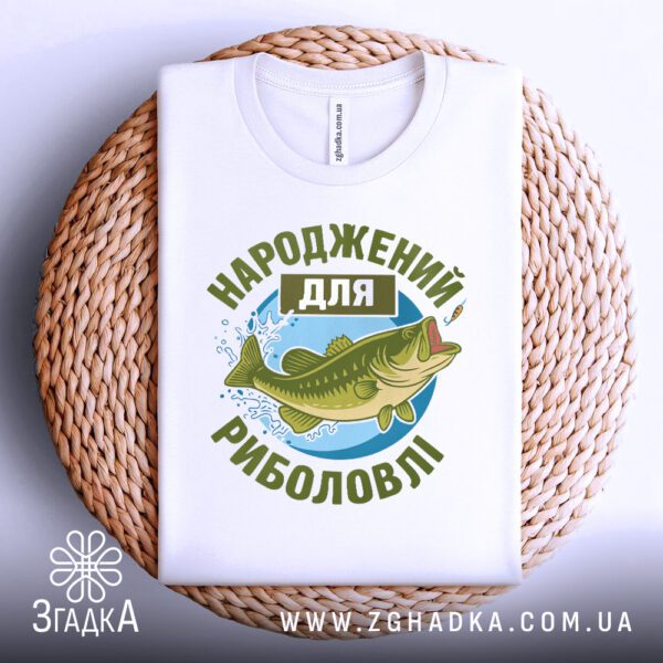 Фронтальний вигляд білої футболки з принтом "Народжений для риболовлі", розташованої на плетеному круглому підносі.