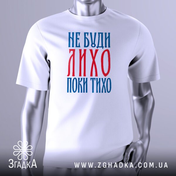 Біла футболка з чорним графічним принтом, що містить український вислів "Не буди лихо, поки тихо". Шрифт стилізований, створюючи ефект ручного написання або стародавнього тексту. Футболка на сірому манекені.