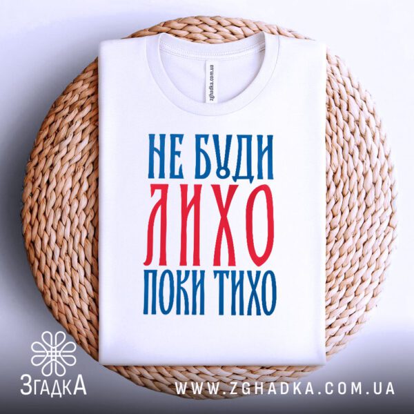 Біла футболка з принтом "не буди лихо", акуратно складена на круглій дерев'яній поверхні.