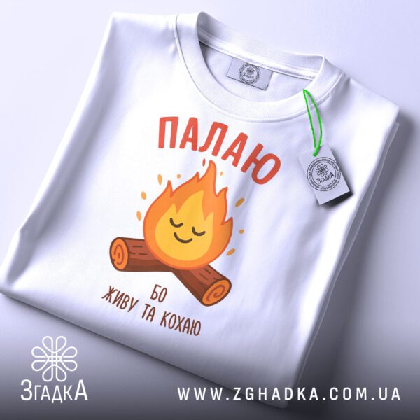 Складена чорна футболка з білим текстовим принтом "Палаю бо живу та кохаю" та етикеткою "Згадка".
