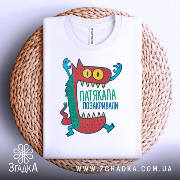 Біла футболка з принтом "Патякала Позакривали", акуратно складена на круглому плетеному підносі.
