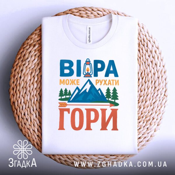 Біла футболка з принтом "рушійна сила віри", акуратно складена на круглій дерев'яній поверхні.