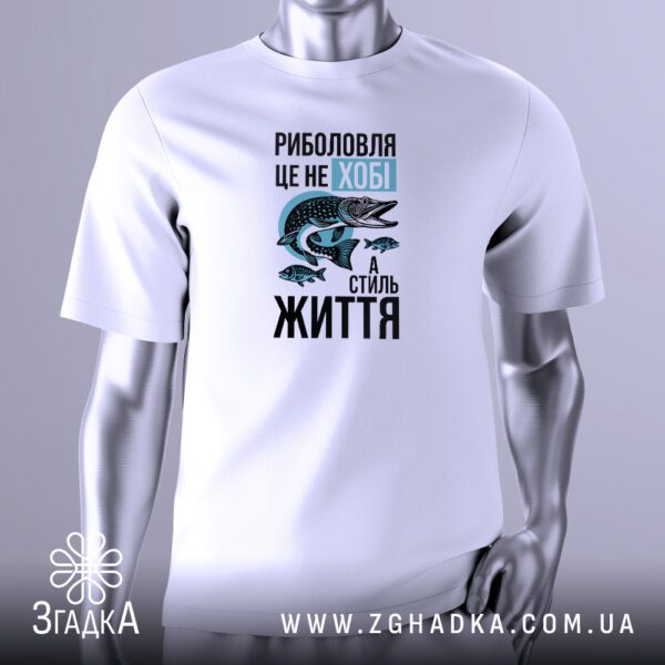 Біла футболка з кольоровим принтом, на якому зображено чоловіка в капелюсі, що сидить на стільці біля води з вудкою, а на гачку – велика риба, над якою напис "Моє хобі – РИБОЛОВЛЯ", розташована на сірому фоні.