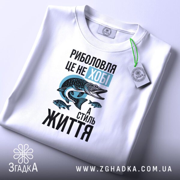 Складена біла футболка з принтом "Моє хобі – РИБОЛОВЛЯ" та етикеткою "Згадка".