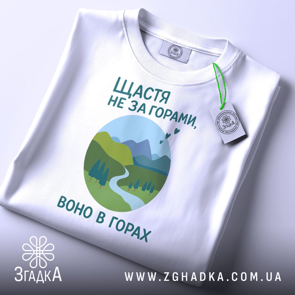 Складена біла футболка з принтом "щастя в горах" та етикеткою "Згадка".
