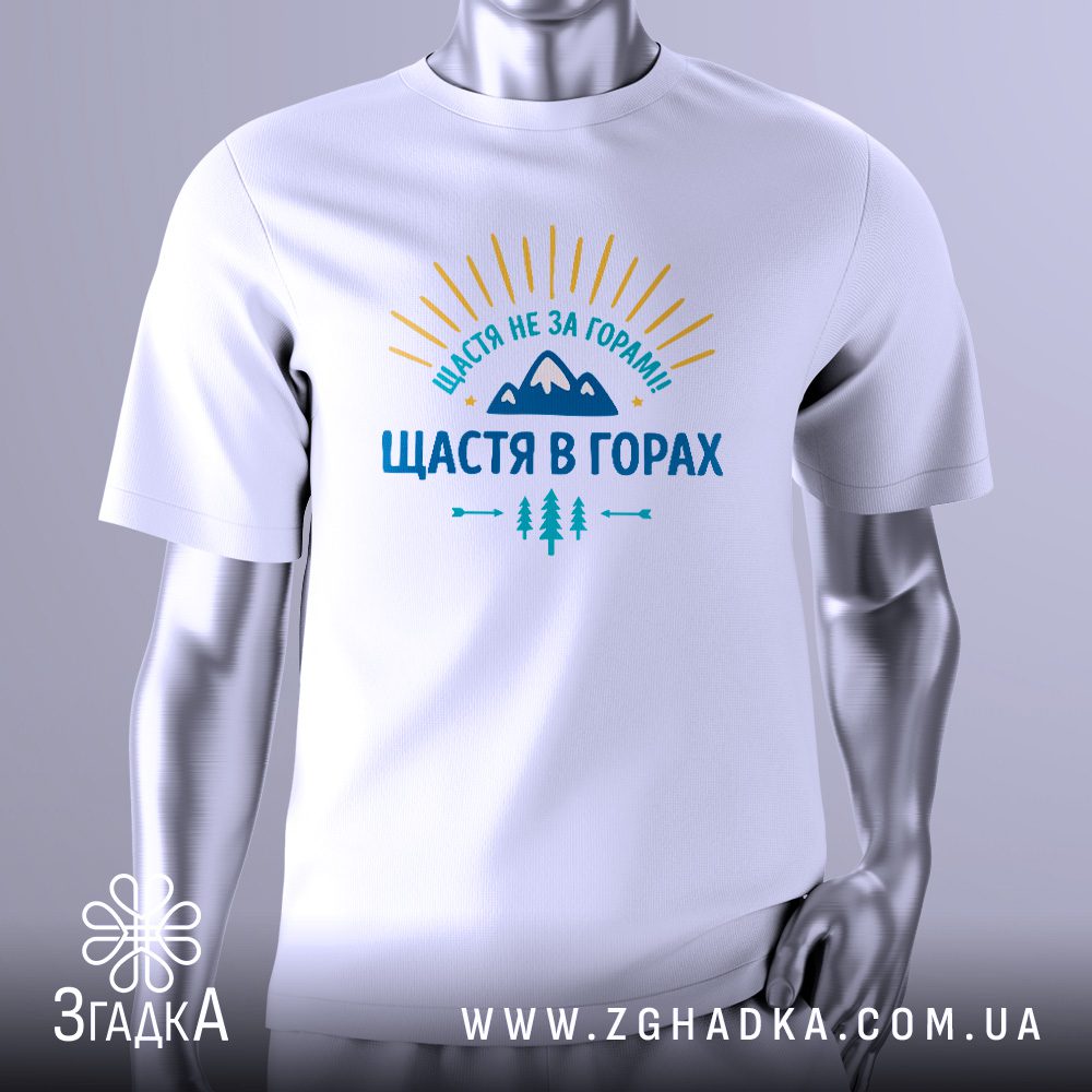 Біла футболка з графічним принтом у центрі. Принт виконаний у темних тонах і зображує стилізовані гірські вершини та промені сонця, що пробиваються крізь них. Над зображенням великими літерами написано фразу: "ЩАСТЯ НЕ ЗА ГОРАМИ - ЩАСТЯ В ГОРАХ". Футболка на сірому манекені.
