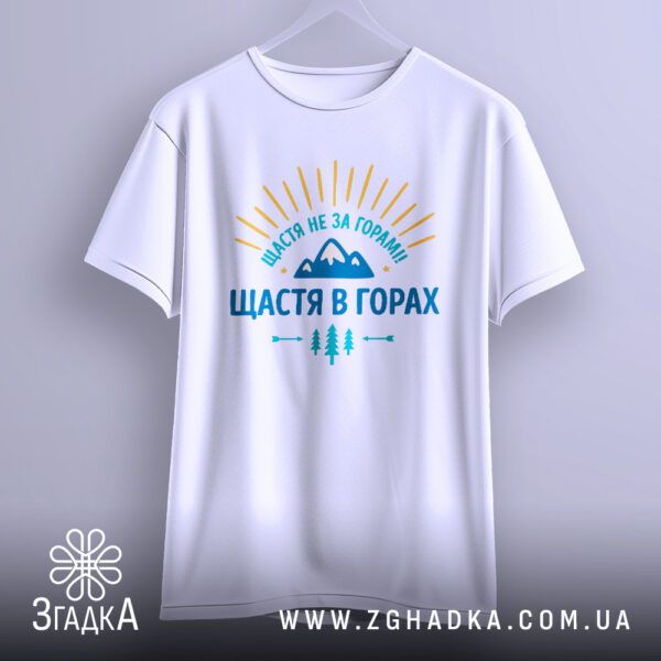 Біла футболка з принтом "щастя в горах", що висить на білих плічках, демонструючи дизайн.