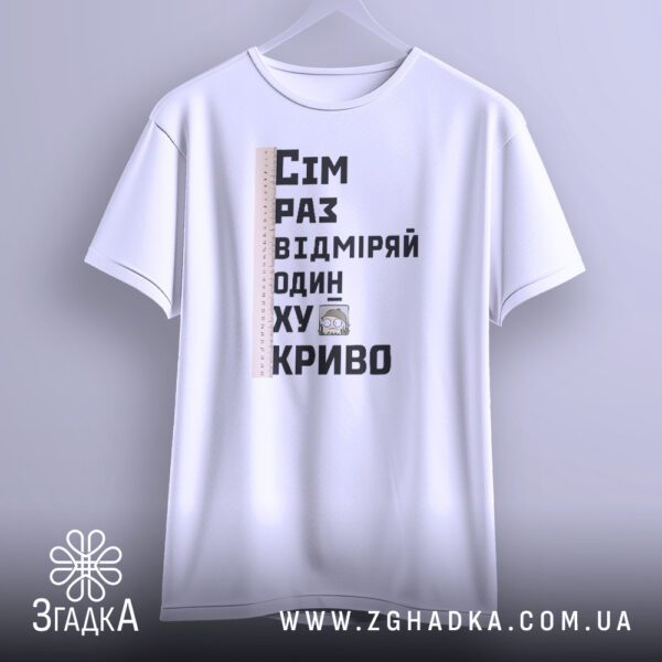 Біла футболка з принтом "сім раз відміряй", що висить на білих плічках, демонструючи дизайн.