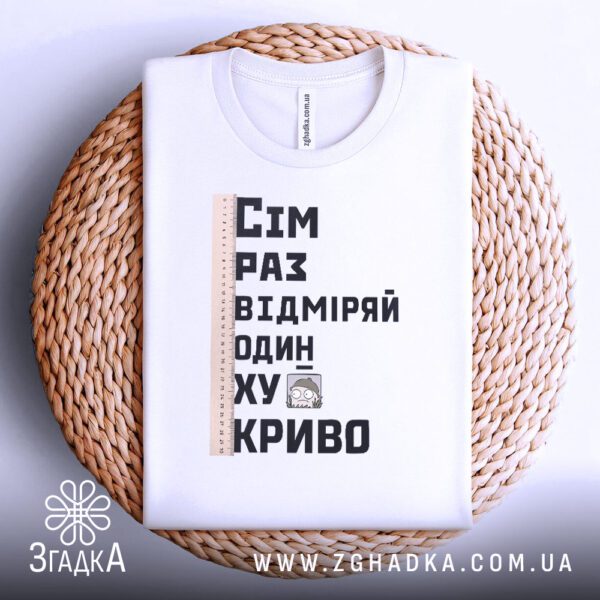 Біла футболка з принтом "сім раз відміряй", акуратно складена на круглій дерев'яній поверхні.