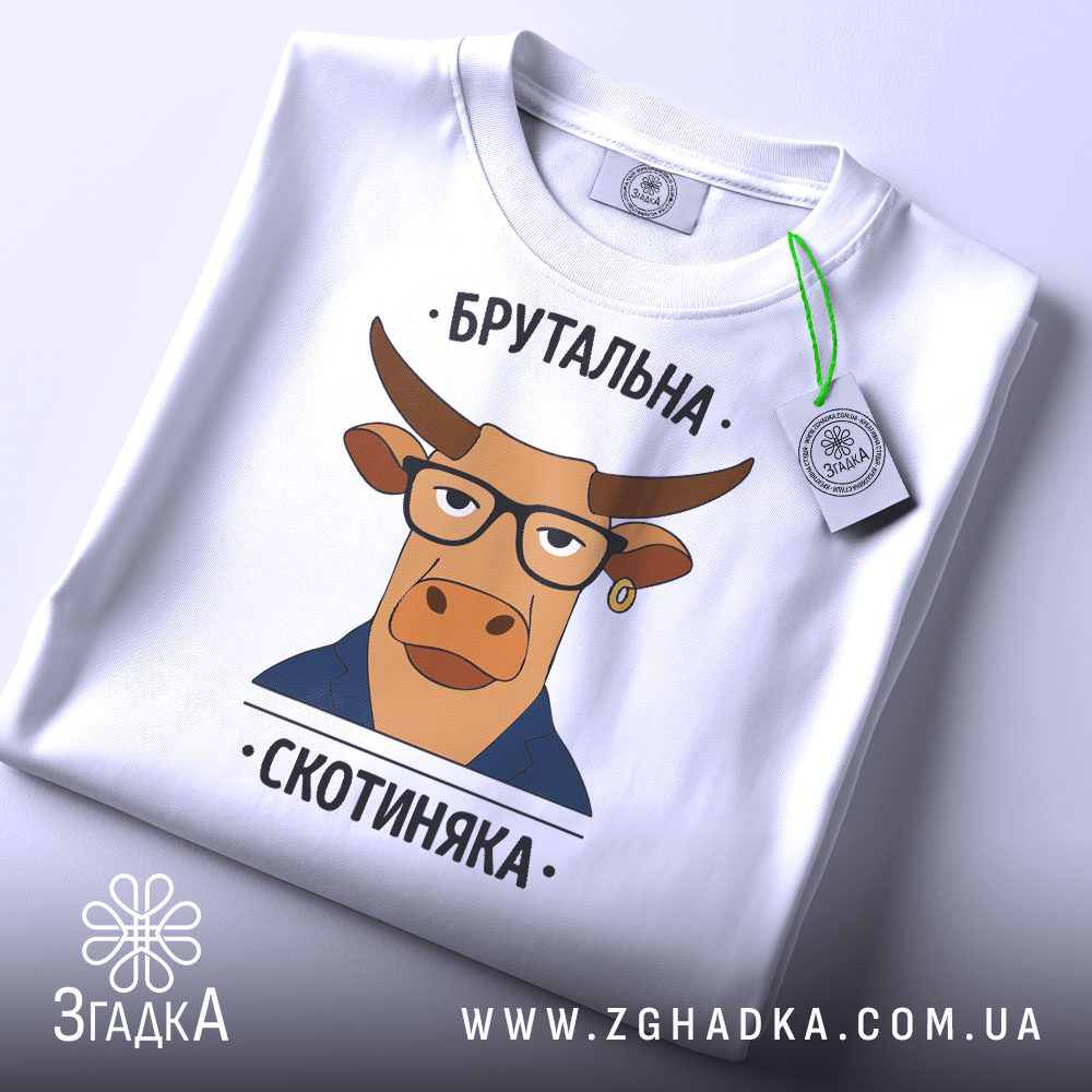 Складена біла футболка з чорним текстовим принтом "Брутальна скотиняка" та етикеткою "Згадка".