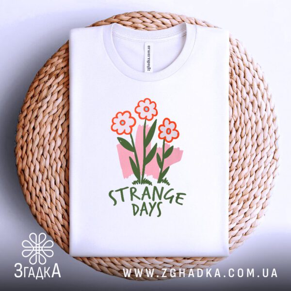 Біла футболка з принтом "strange days квіти", акуратно складена на круглій дерев'яній поверхні.