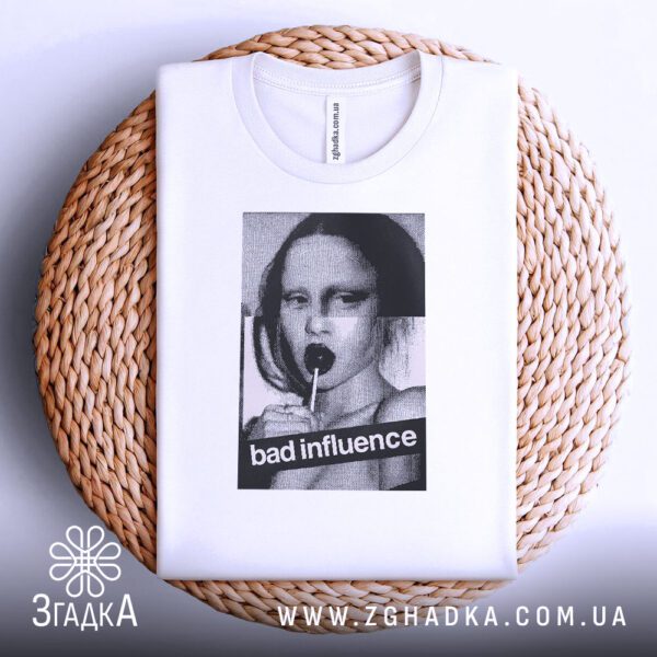 Біла футболка з принтом "bad influence", акуратно складена на круглій дерев'яній поверхні.