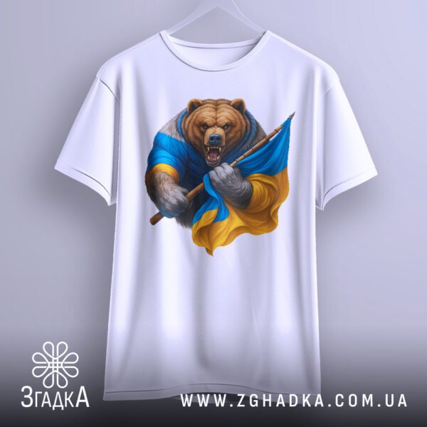 Біла футболка з принтом "ведмідь з прапором", що висить на білих плічках, демонструючи дизайн.