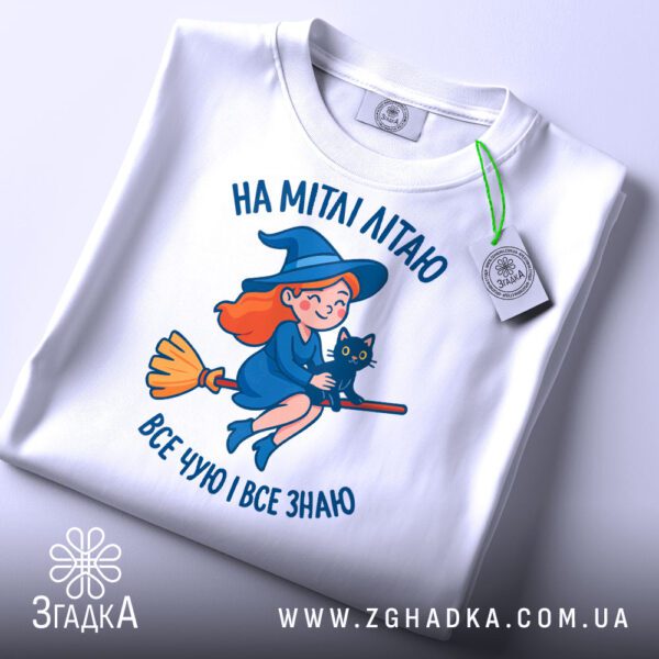 Складена біла футболка з принтом відьмочки на мітлі та етикеткою "Згадка".